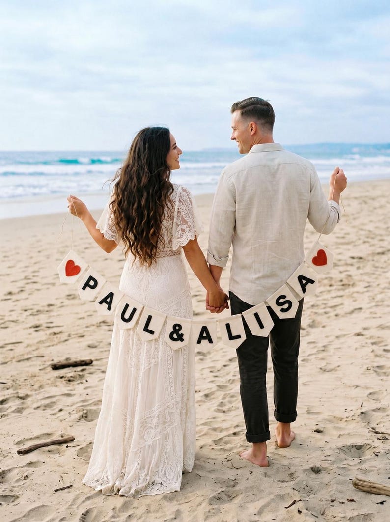 May include: A cream-coloured fabric banner featuring the text PAUL & ALISA in black capital letters, flanked by two red heart icons on each end. The banner is suspended by a thin string and held horizontally between two individuals on a sandy beach.