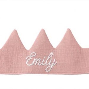 K&ouml;nnte beinhalten: Ein hellrosa Stoffkrone mit drei Zacken und dem Namen "Emily" in wei&szlig;er Schrift gestickt. Die Krone hat rosa B&auml;nder zum Binden. Entworfen f&uuml;r Kinder.