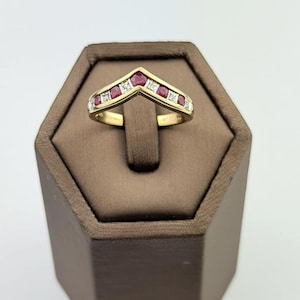 May include: A gold chevron-shaped ring featuring a series of alternating round red rubies and small clear diamonds set along the top band. The ring has a polished yellow gold finish and a V-shaped design that tapers toward the center point.