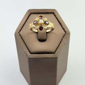 May include: A gold ring featuring a central white cabochon stone surrounded by four oval dark red garnet stones and four small white seed pearls. The band has a textured, leaf-like pattern on the shoulders, creating a cluster design.