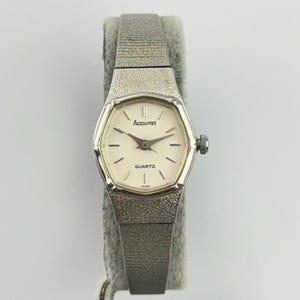 Peut inclure: Une montre à quartz Accurist argentée avec un boîtier hexagonal et un bracelet en métal texturé. Le cadran blanc cassé présente de fins index noirs, des aiguilles noires et le texte Accurist et QUARTZ imprimé en noir sur le cadran.
