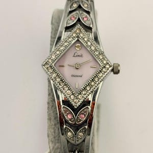 Könnte beinhalten: Eine silberfarbene Analoguhr mit rautenförmigem Zifferblatt in Hellrosa, beschriftet mit Limit und Diamond. Die Lünette und das Armband sind mit klaren Kristallen und kleinen rosa Akzenten in einem blattartigen Muster verziert.