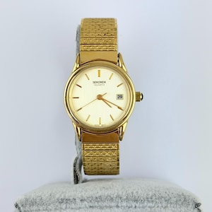 May include: A gold-tone Sekonda Quartz wristwatch featuring a round face with a light champagne dial, gold-tone hour markers, and a date window at the three o'clock position. The watch includes a textured, flexible gold-tone metal expansion band.