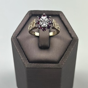 May include: A gold-toned ring with a floral design. The center features a large, clear stone surrounded by smaller, deep red stones. The band has decorative details on the sides, and the ring is displayed on a brown jewelry stand.