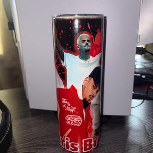 May include: A stainless steel tumbler featuring images of Chris Brown. The tumbler has a black top and a red and black design with the text "Chris Br". The tumbler is cylindrical and appears to be approximately 8 inches tall.