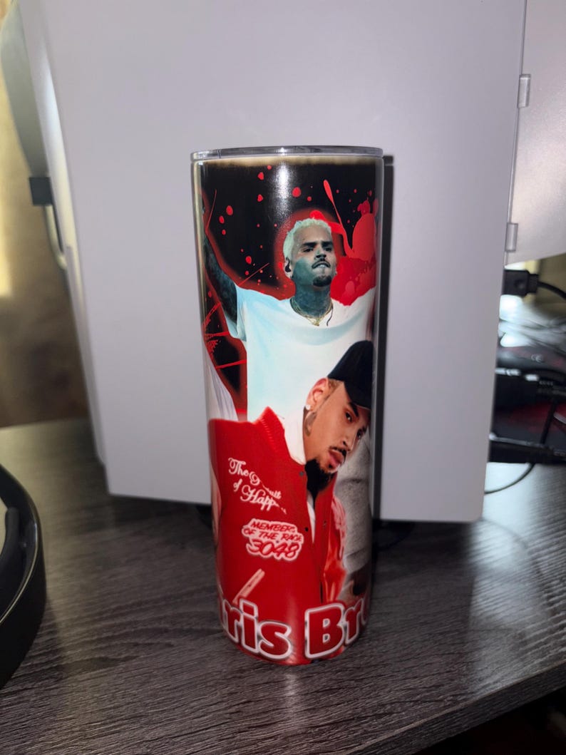May include: A stainless steel tumbler featuring images of Chris Brown. The tumbler has a black top and a red and black design with the text "Chris Br". The tumbler is cylindrical and appears to be approximately 8 inches tall.