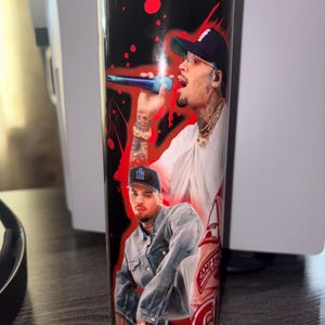 May include: A black and red tumbler featuring two images of a man. The tumbler has a black background with red splatter designs. One image shows the man singing into a microphone, and the other shows him in a denim jacket.