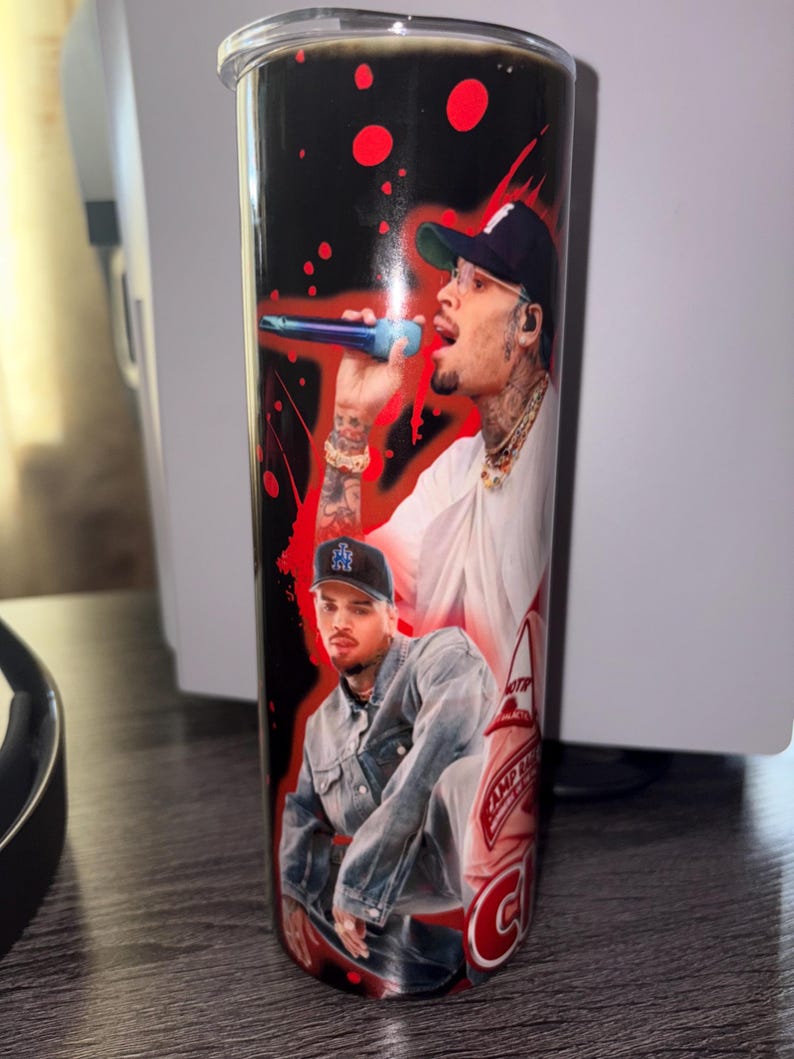 May include: A black and red tumbler featuring two images of a man. The tumbler has a black background with red splatter designs. One image shows the man singing into a microphone, and the other shows him in a denim jacket.
