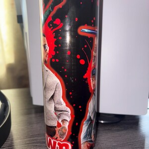 May include: A cylindrical candle with a black background and red splatter design. The candle features two images of a person wearing sunglasses and casual clothing. The word "WN" is visible at the bottom of the candle.