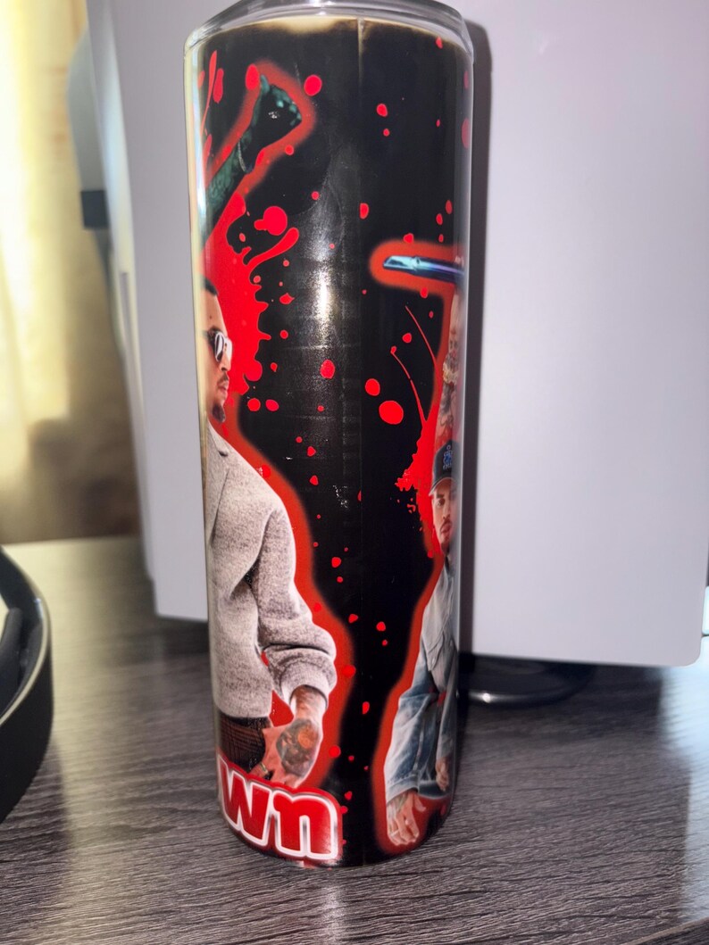 May include: A cylindrical candle with a black background and red splatter design. The candle features two images of a person wearing sunglasses and casual clothing. The word "WN" is visible at the bottom of the candle.