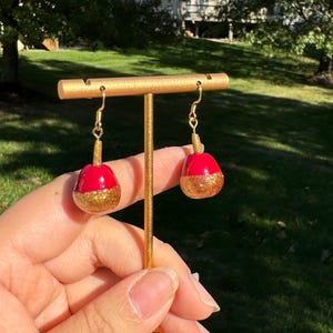 May include: A pair of dangle earrings with red and gold accents. The earrings are shaped like apples with a gold glitter finish.