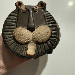 Puede incluir: Figura de león de cerámica en tonos marrón y beige. El león tiene un rostro detallado con rasgos texturizados, como una nariz prominente, mejillas redondas y cejas fruncidas. La melena se representa con líneas horizontales.