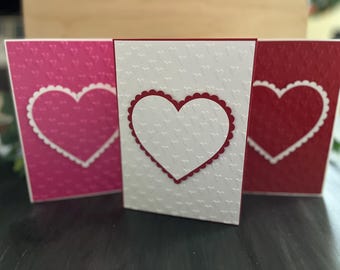 May include: Three Valentine's Day cards with different colored backgrounds. The cards are decorated with white hearts with scalloped edges. The hearts are on a background of pink, white, and red.