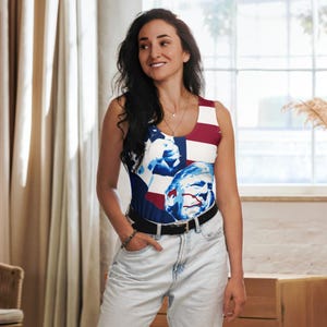 K&ouml;nnte beinhalten: Eine Frau tr&auml;gt ein &auml;rmelloses Tanktop mit einem rot-wei&szlig;-blauen amerikanischen Flaggenmuster. Das Tanktop zeigt eine Grafik mit dem Gesicht und der Hand einer Person. Die Frau tr&auml;gt eine blaue Jeans und einen braunen G&uuml;rtel.