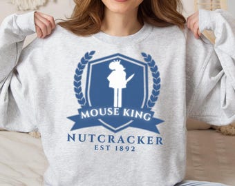 May include: Light gray sweatshirt featuring a blue and white graphic. The graphic includes the words "MOUSE KING" and "NUTCRACKER EST 1892" with a silhouette of a figure. The design is framed by a laurel wreath.