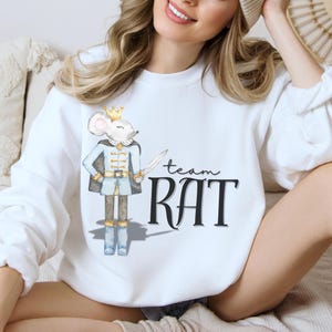 May include: White sweatshirt featuring a watercolor illustration of a mouse in a blue and gold uniform, holding a sword. The text "team RAT" is printed in black script and block letters. The sweatshirt is a casual, comfortable style.