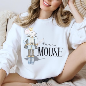 Könnte beinhalten: Weißes Sweatshirt mit einem Aquarell-Motiv einer Maus in blau-goldener Kleidung, die ein Schwert hält. Der Text "team MOUSE" ist in schwarzer Schrift und Blockbuchstaben gedruckt. Das Sweatshirt hat einen Rundhalsausschnitt.