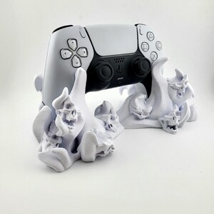 Może przedstawiać: Biało-czarny kontroler PlayStation 5 spoczywa na białym stojaku z motywem czaszki. Stojak posiada wiele czaszek i szczegółowy, teksturowany design. Kontroler ma elegancki, nowoczesny design z czarnymi joystickami i przyciskami.