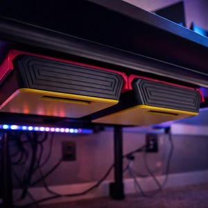 May include: Two black and red under-desk storage drawers with yellow accents are mounted under a black desk. The drawers have a textured design and are illuminated by colorful LED lights. The desk is supported by black legs.