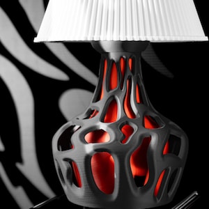 May include: A modern table lamp with a white pleated lampshade. The lamp base is dark gray with an open, organic design revealing a bright red interior. The background features a black and white zebra stripe pattern.