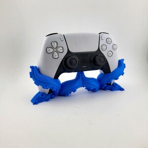May include: A white and black PlayStation 5 controller is held by a blue octopus-shaped stand. The controller has the standard button layout and a central touchpad. The stand's tentacles wrap around the controller, providing support.