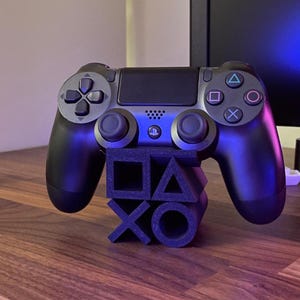 Puede incluir: Un mando negro de PlayStation 4 apoyado en un soporte negro con los símbolos de PlayStation. El mando tiene un panel táctil, botones direccionales y joysticks. El soporte está hecho de formas apiladas.