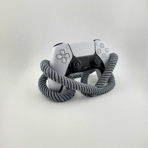Könnte beinhalten: Ein weiß-schwarzer PlayStation 5-Controller wird von einer grauen, seilartigen Struktur umschlossen. Der Controller verfügt über ein zentrales Touchpad, Richtungstasten und analoge Sticks. Das graue Seil sorgt für einen textlichen Kontrast.