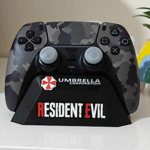 May include: A camouflage-patterned PlayStation 5 controller resting on a black stand. The stand features the "Umbrella Corporation" logo and "Resident Evil" text in red and white.