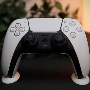 Puede incluir: Un mando inalámbrico DualSense blanco y negro para PlayStation 5. El mando tiene una sección central negra con dos joysticks analógicos negros y un logotipo de PlayStation. Los lados blancos presentan la disposición estándar de botones.