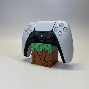 Könnte beinhalten: Ein weiß-schwarzer PlayStation 5-Controller liegt auf einem braun-grünen, pixeligen Block. Der Controller hat ein schlankes Design mit verschiedenen Tasten und Joysticks. Der Block ähnelt einem Minecraft-Stil.