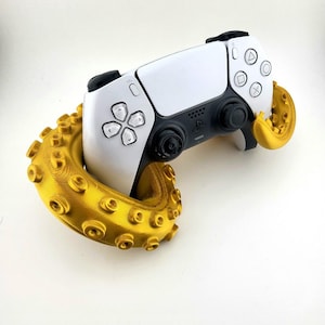 May include: A white and black PlayStation 5 controller is held by a gold-colored tentacle-shaped stand. The controller has black buttons and directional pads. The stand has a textured surface with suction cup details.