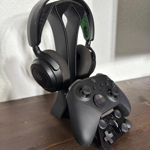 May include: Black gaming headset and controller resting on a black stand. The headset has a black frame with gray earcups and green accents. The controller is black with multiple buttons and joysticks, designed for video game play.