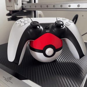 Puede incluir: Un mando blanco y negro de PlayStation 5 con una Poké Ball roja y blanca adjunta. El mando está sobre una superficie negra con un patrón de fibra de carbono. La imagen está bien iluminada y muestra los botones y joysticks.