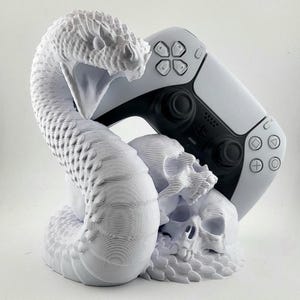 May include: White 3D-printed gaming controller stand featuring a snake coiled around two skulls. The stand is designed to hold a white and black gaming controller. The snake has detailed scales and an open mouth.