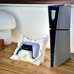 May include: A white and black game controller rests on a white satin pillow. A silver and black game console stands upright to the right. The console has a black center with a white accent and a blue light. The objects are on a wooden surface.