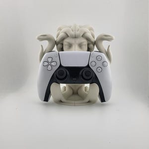 May include: A white and black PlayStation 5 controller rests in a light beige Medusa head-shaped stand. The controller features black buttons and directional pads. The stand has two snakes and detailed facial features.