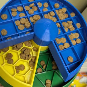 May include: A multi-colored pet food puzzle in blue, yellow, and green. The puzzle is filled with brown kibble and has a rotating center. The design features various compartments and shapes to dispense food.
