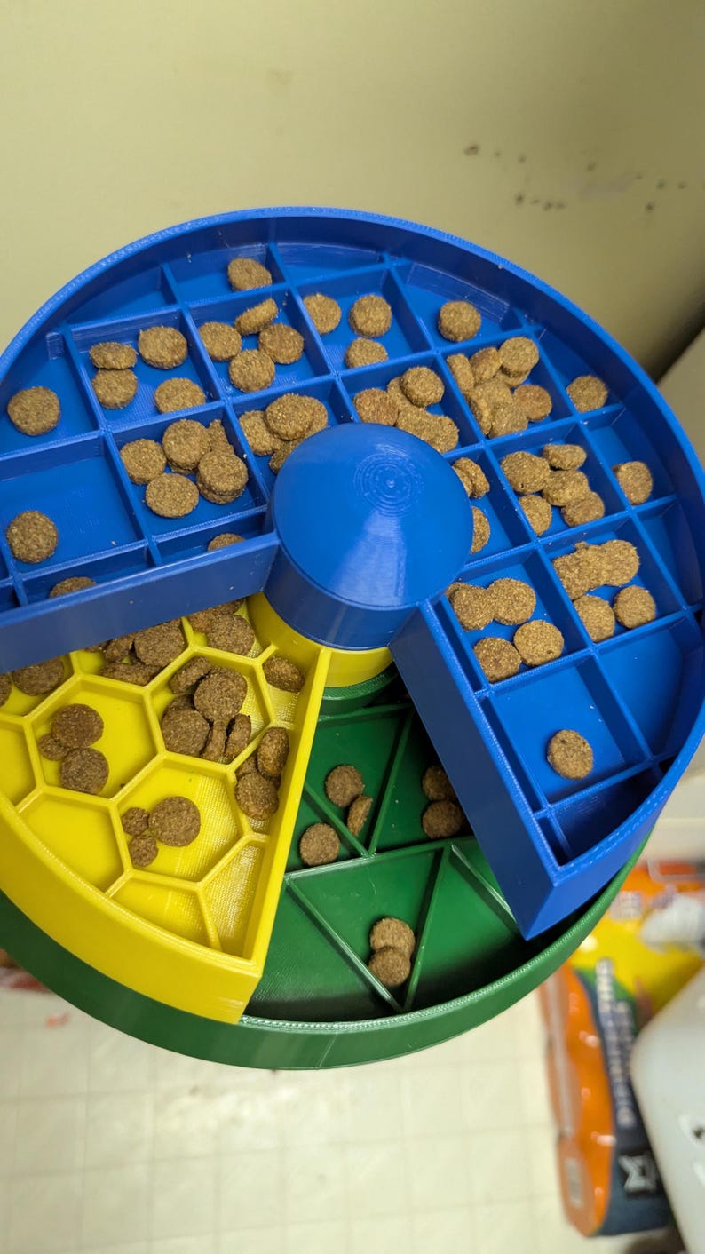 May include: A multi-colored pet food puzzle in blue, yellow, and green. The puzzle is filled with brown kibble and has a rotating center. The design features various compartments and shapes to dispense food.