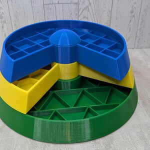 May include: A multi-colored, tiered storage container. The base is green, the middle section is yellow, and the top section is blue. The top section has a grid-like pattern. A blue, rounded top is in the center.