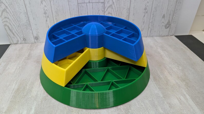 May include: A multi-colored, tiered storage container. The base is green, the middle section is yellow, and the top section is blue. The top section has a grid-like pattern. A blue, rounded top is in the center.