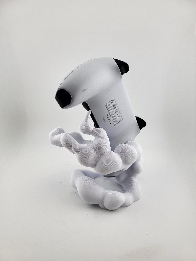 Op de afbeelding: Een wit en zwarte gamecontroller op een witte, wolkvormige standaard. De controller is omhoog gekanteld en wordt ondersteund door de standaard. De standaard heeft een gestructureerd, organisch ontwerp.