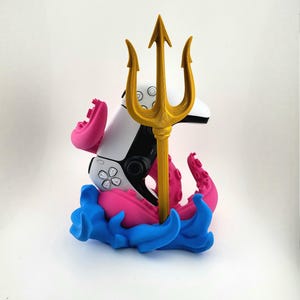 Könnte beinhalten: Ein weiß-schwarzer Gamecontroller ist mit einem goldenen Dreizack, rosa Tentakeln und blauen Wellen verziert. Der Controller steht im Mittelpunkt, mit dem Dreizack, der aus der Mitte ragt. Die Basis zeigt ein lebendiges blaues Wellendesign.