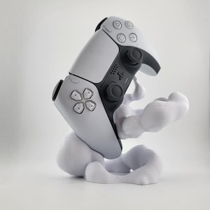 May include: A white and black PlayStation 5 DualSense controller resting on a white, cloud-shaped stand. The controller has a symmetrical design with buttons and joysticks. The stand has a smooth, flowing form.