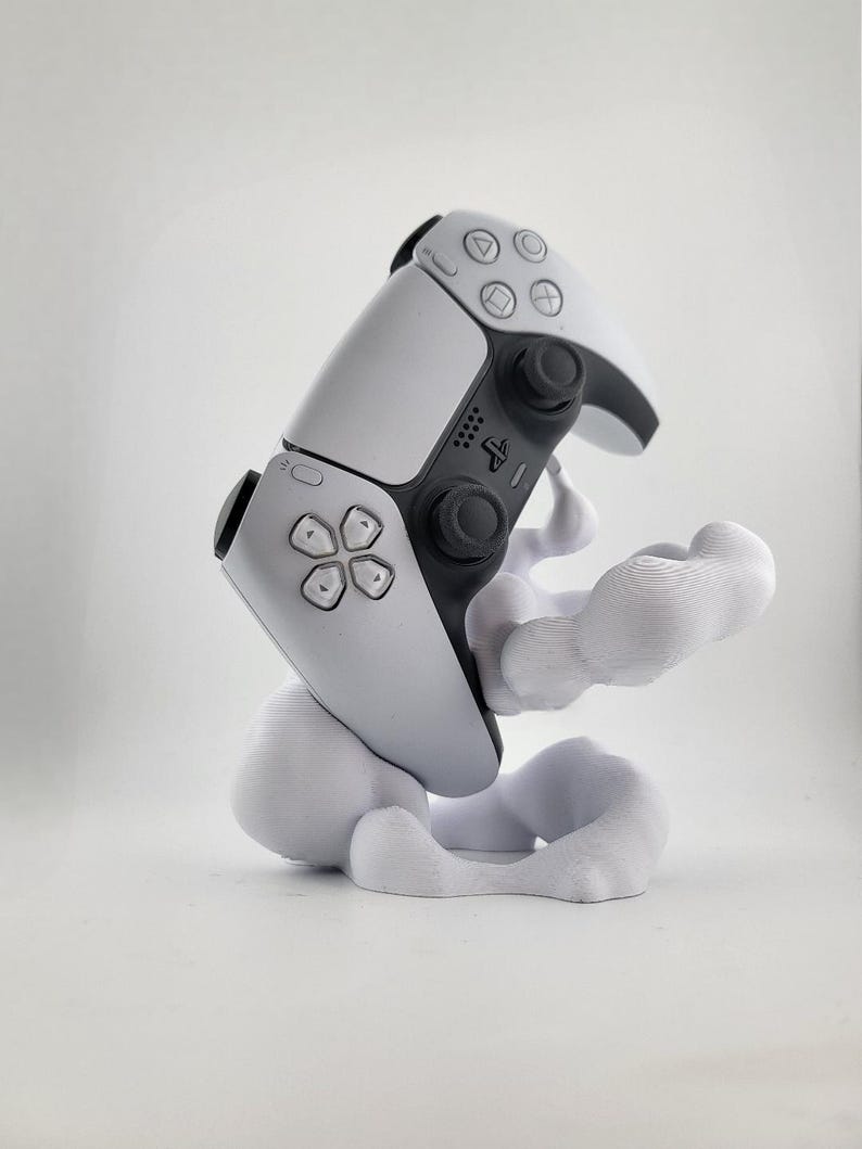 Op de afbeelding: Een wit en zwarte PlayStation 5 DualSense controller rustend op een witte, wolkvormige standaard. De controller heeft een symmetrisch ontwerp met knoppen en joysticks. De standaard heeft een gladde, vloeiende vorm.