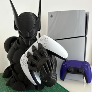 May include: A black and white robot-themed controller holder with a white PlayStation 5 controller in its hands. A white PlayStation 5 console and a purple controller are also visible.