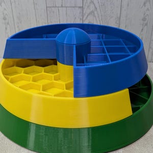 May include: A multi-tiered, 3D-printed organizer in primary colors. The base is green, supporting a yellow layer with a honeycomb pattern. A blue section with compartments and a central dome sits atop the yellow layer. The design is geometric and functional.