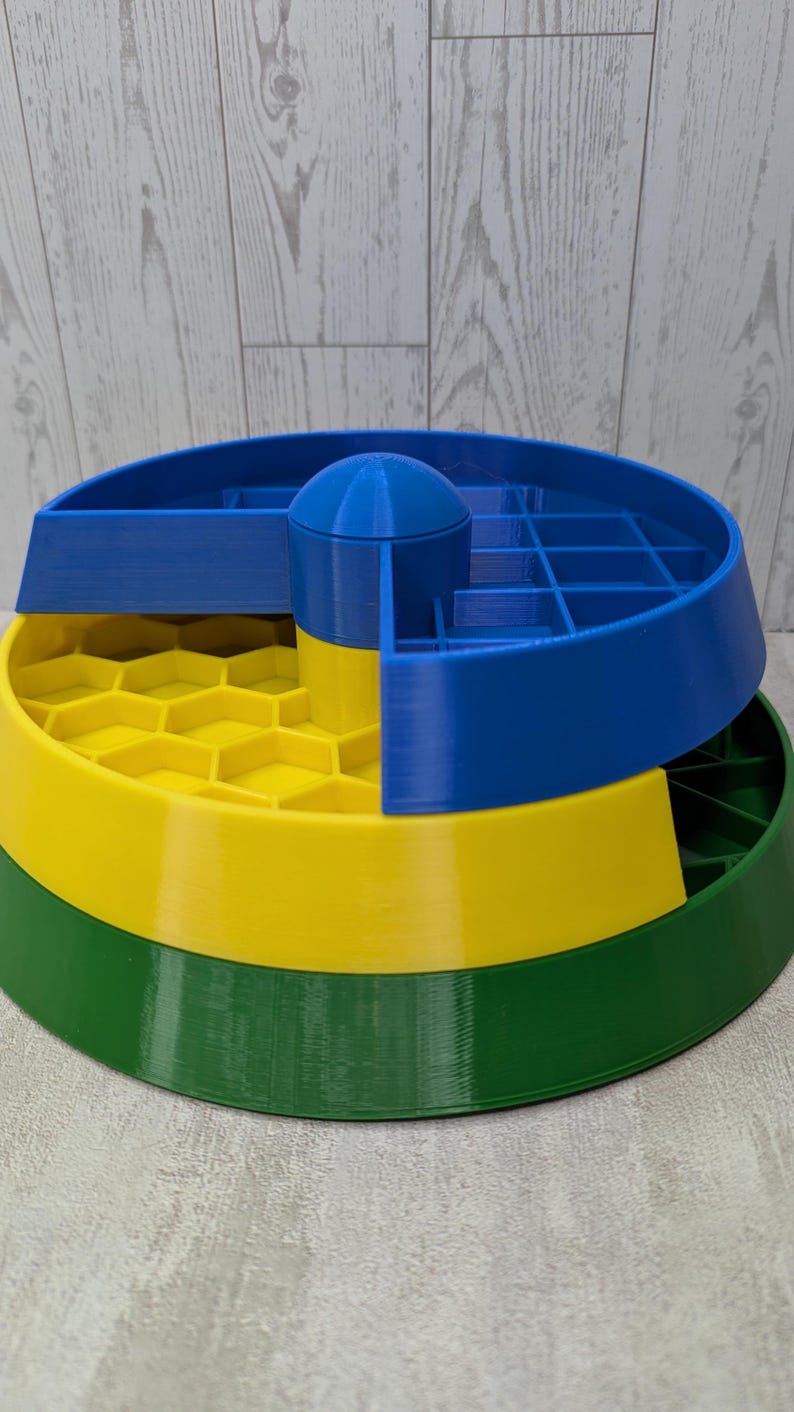 May include: A multi-tiered, 3D-printed organizer in primary colors. The base is green, supporting a yellow layer with a honeycomb pattern. A blue section with compartments and a central dome sits atop the yellow layer. The design is geometric and functional.