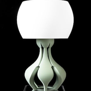 Puede incluir: Lámpara de mesa moderna con una pantalla blanca y redondeada. La base es de color verde claro con un diseño calado único. La lámpara está sobre un fondo negro, resaltando su estética contemporánea. Altura aproximada: 40 cm.