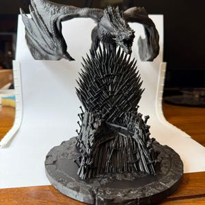 May include: A black 3D-printed sculpture of a dragon perched atop the Iron Throne. The dragon has outstretched wings and a detailed, textured appearance. The throne is made of numerous swords and sits on a circular base.