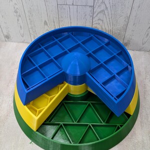 May include: A multi-tiered, 3D-printed organizer in primary colors. The base is green, supporting a yellow layer with hexagonal compartments. A blue top layer features a grid of square compartments. The organizer is designed for small items.
